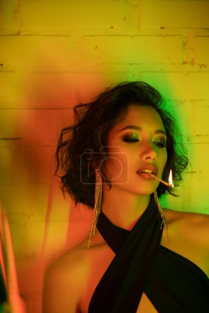 Asian woman in earrings and dress holding match with fire in lips in night club with neon light