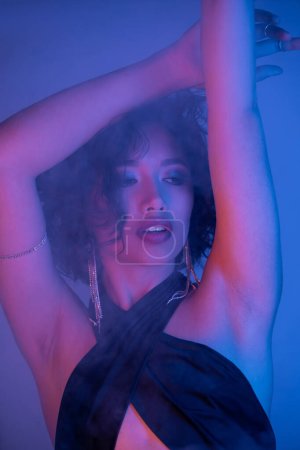 Young asian woman in earrings and dress dancing in neon light and smoke in night club
