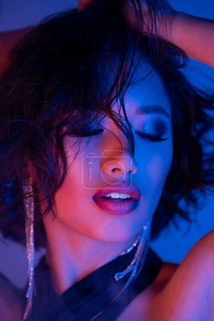 Stilvolle asiatische Frau mit Frisur und Make-up posiert mit geschlossenen Augen im Neonlicht im Nachtclub