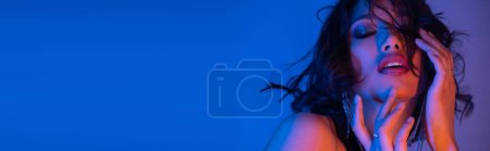 Glamours young asian woman touching face while posing in neon light in night club, banner