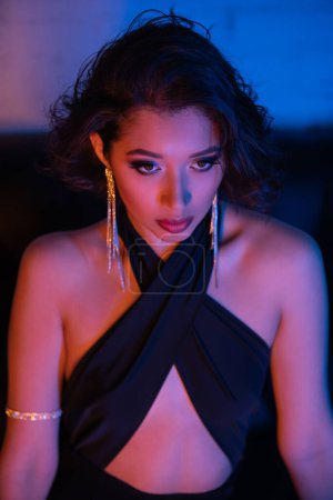 Confident asian woman in stylish dress and earrings sitting on couch in neon light in night club