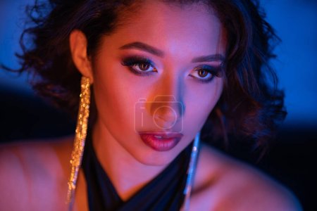 Retrato de mujer asiática con estilo con maquillaje mirando hacia otro lado en luz de neón en club nocturno borroso