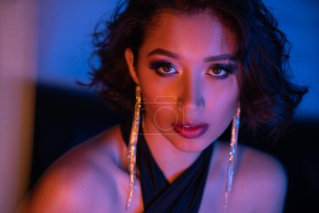 Porträt einer stilvollen asiatischen Frau, die auf einer verschwommenen Couch im Neonlicht eines Nachtclubs in die Kamera blickt