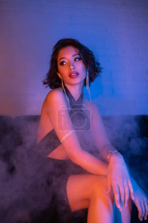 Modische junge asiatische Frau im Kleid sitzt in Rauch und Neonlicht auf der Couch in einem Nachtclub