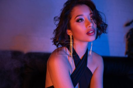 Elegant young asian woman in evening wear standing in pink and blue neon light in night club