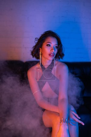 Elegante junge asiatische Frau in Abendgarderobe sitzt in Rauch und Neonlicht auf der Couch im Nachtclub