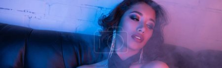 Fashionable asian woman with makeup sitting on couch with colorful neon light in night club, banner