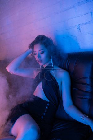 Sexy asian woman in dress touching hair and sitting on couch near smoke and neon light in night club