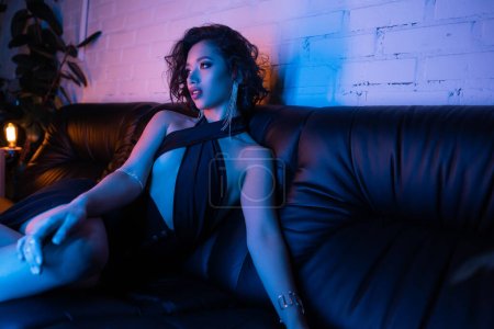 Stilvolle und sexy asiatische Frau im Abendkleid sitzt auf der Couch in buntem Neonlicht in Nachtclub