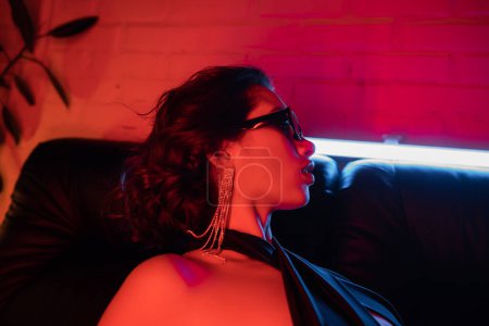 Photo for Side view of sexy asian woman in sunglasses and earring posing on couch in neon light in night club - Royalty Free Image