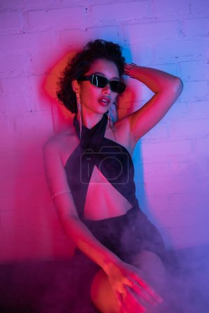 Stilvolle asiatische Frau mit Sonnenbrille, die Haare berührt und in Rauch und Neonlicht in einem Nachtclub sitzt