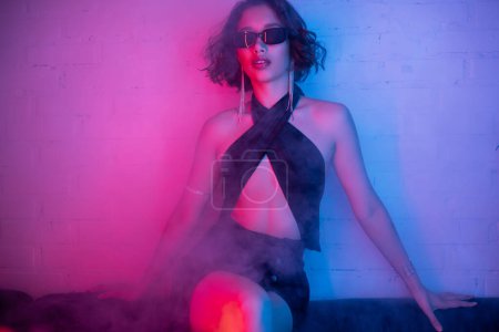 Modische asiatische Frau in sexy Kleid und Sonnenbrille posiert in Rauch und Neonlicht in Nachtclub