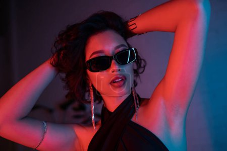 Photo for Sexy asian woman in sunglasses and bracelets touching head in neon light in night club - Royalty Free Image