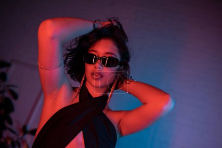 Trendy asian woman in sunglasses and dress touching head in colorful neon lighting in night club