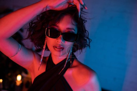 Fashionable asian woman in sunglasses and earrings standing in vivid neon light in night club