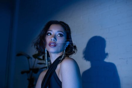Téléchargez les photos : Jolie femme asiatique avec maquillage et coiffure regardant loin tout en se tenant dans la lumière au néon dans la boîte de nuit - en image libre de droit