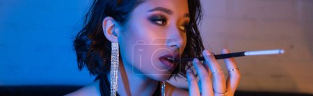 Photo for Trendy asian woman with visage holding cigarette in mouthpiece in night club with neon light, banner - Royalty Free Image