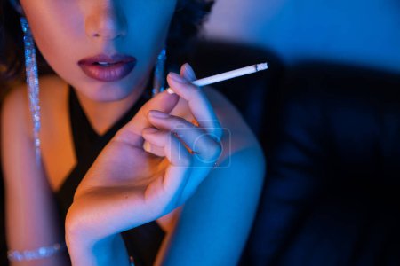 Ausgeschnittene Ansicht einer stilvollen Frau im Kleid mit Zigarette, während sie im Neonlicht in einem Nachtclub sitzt