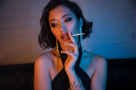 Sexy junge asiatische Frau im Kleid raucht Zigarette und verbringt Zeit in einem Nachtclub mit Neonlicht
