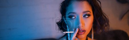 Fashionable asian woman with makeup and hairstyle smoking cigarette in night club, banner