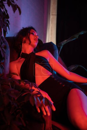 Sexy asian woman in evening dress sitting on armchair near plants in night club with neon lighting