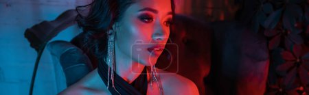 Stylish young asian woman with makeup standing near plants in neon light in night club, banner
