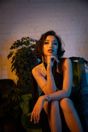 Stilvolle junge asiatische Frau mit Make-up und Frisur sitzt auf Sessel in Nachtclub