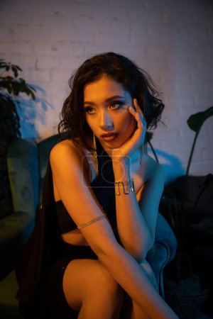 Stylish asian woman holding hand near cheek while sitting on armchair in night club with lighting