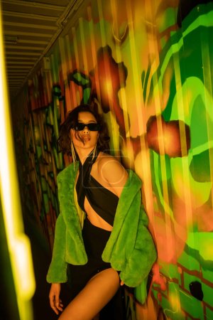 Sexy asiatische Frau mit Sonnenbrille und Kunstpelzjacke steht in der Nähe von Graffiti in Nachtclub