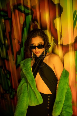 Sexy and pretty asian woman in sunglasses and fake fur jacket near graffiti on wall in night club