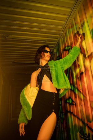 Low angle view of asian woman in sunglasses and fake fur jacket standing near graffiti in night club