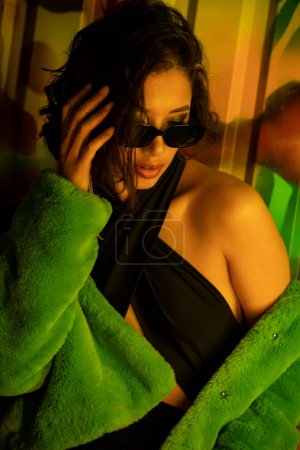 Stylish asian woman in sunglasses touching hair while standing near graffiti on wall in night club
