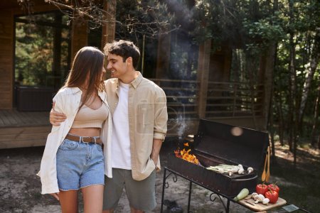 homme heureux étreignant petite amie près du barbecue grill, romance d'été, couple, maison de vacances