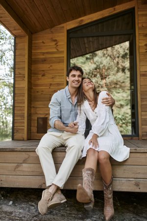 Mann umarmt fröhliche Frau im Sitzen auf Veranda, glückliches Paar, Ferienhaus, Sommerromantik