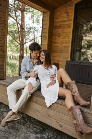 romantischer Mann umarmt fröhliche Frau, während sie zusammen auf der Veranda sitzt, Paar, Ferienhaus, Urlaub