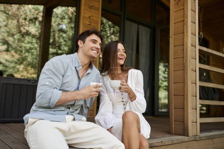 happy couple holding cups of morning coffee, man and woman laughing on porch of vacation house