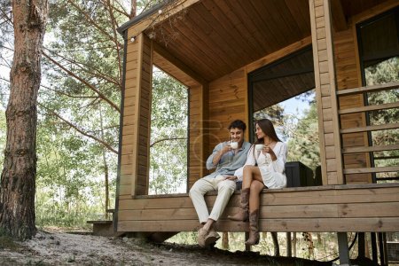 happy couple holding cups of coffee, man and woman smiling on porch of vacation house in forest