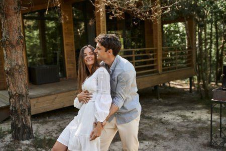 happy man hugging woman near vacation house in green forest, summer and romance, couple in love