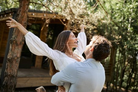 Téléchargez les photos : Homme heureux soulevant femme avec les mains tendues, maison de vacances près de la forêt, romance et amour - en image libre de droit