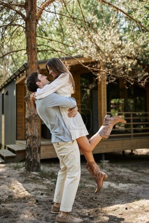 happy man lifting and hugging woman in vacation house, forest, look at each other, romance and love