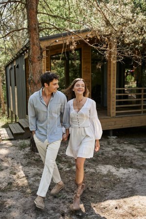 happy woman holding hands and walking with smiling boyfriend, couple near summer house, romance