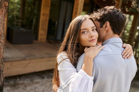 Smiling young woman in sundress hugging boyfriend and looking at camera near vacation house outdoors