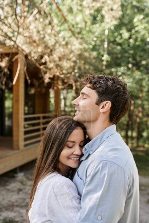 Lächelndes Paar mit geschlossenen Augen umarmt und steht in der Nähe von Ferienhaus im Hintergrund im Freien