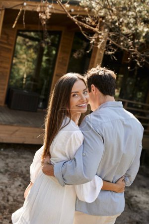 Smiling brunette woman in sundress hugging boyfriend and looking at camera near vacation house