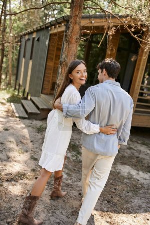 Trendy and smiling woman in sundress hugging boyfriend and looking at camera near vacation house