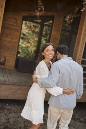 Joyful young woman in sundress hugging boyfriend and looking at camera near vacation house outdoors
