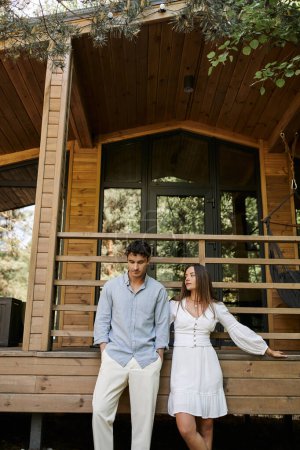 Téléchargez les photos : Femme à la mode en robe de soleil debout près de petit ami brunette et maison de vacances en arrière-plan - en image libre de droit