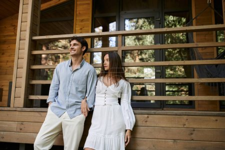 Téléchargez les photos : Homme souriant en vêtements décontractés debout près de petite amie en robe de soleil et maison de vacances à l'arrière-plan - en image libre de droit