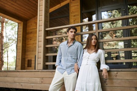 Trendy romantic couple in casual clothes looking away near vacation house outdoors at background