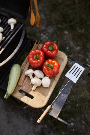 Vista superior de verduras frescas en la tabla de cortar cerca de la barbacoa durante el picnic al aire libre, comida y naturaleza